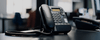 A black telephone has been placed on a black desk. The background is an office with desks and chairs.
