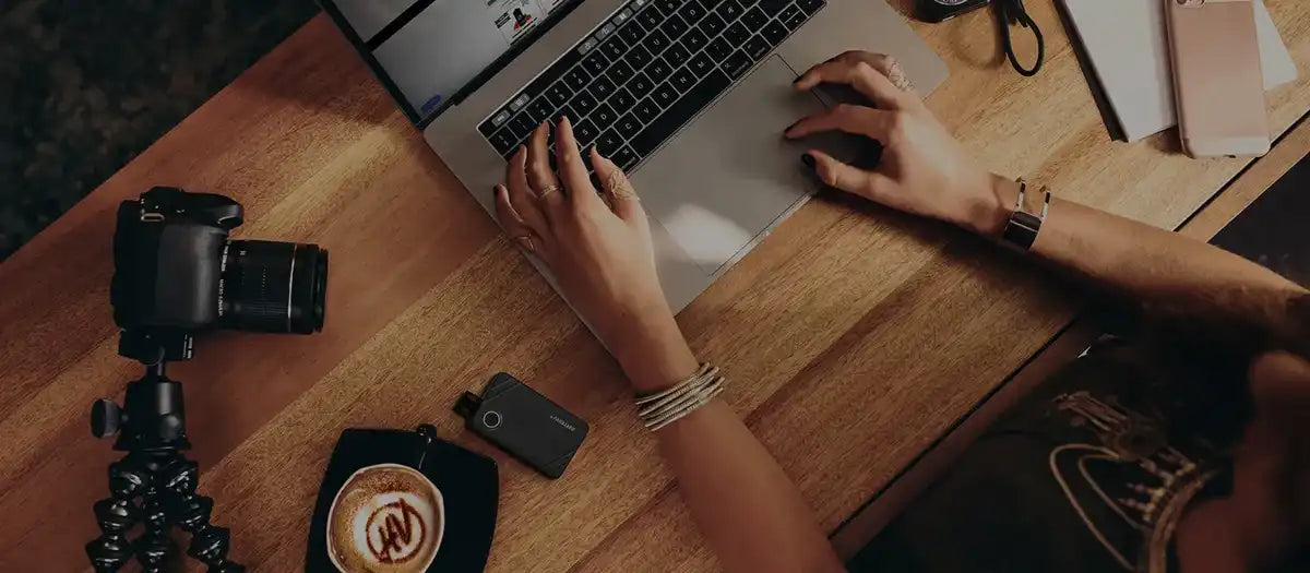 A birds eye view of someone working on a laptop, with a camera, coffee & vape device to their left.