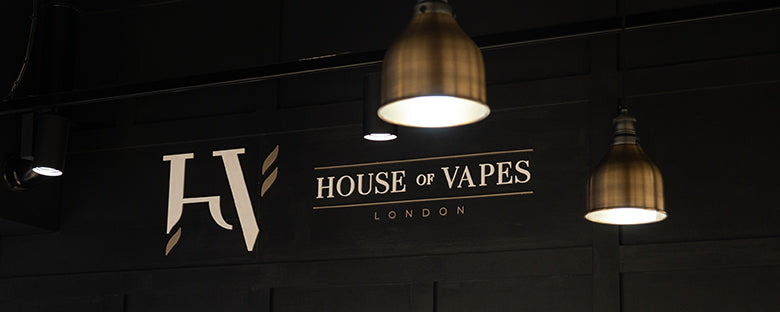The House of Vapes - London logo is shown in gold painted on a black wall. Large gold lights are also shining down onto the logo.